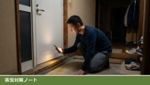 夜の玄関で、男性がスマホのライトでドアの下端と床の境目を照らして、すき間の具合を確かめている様子
